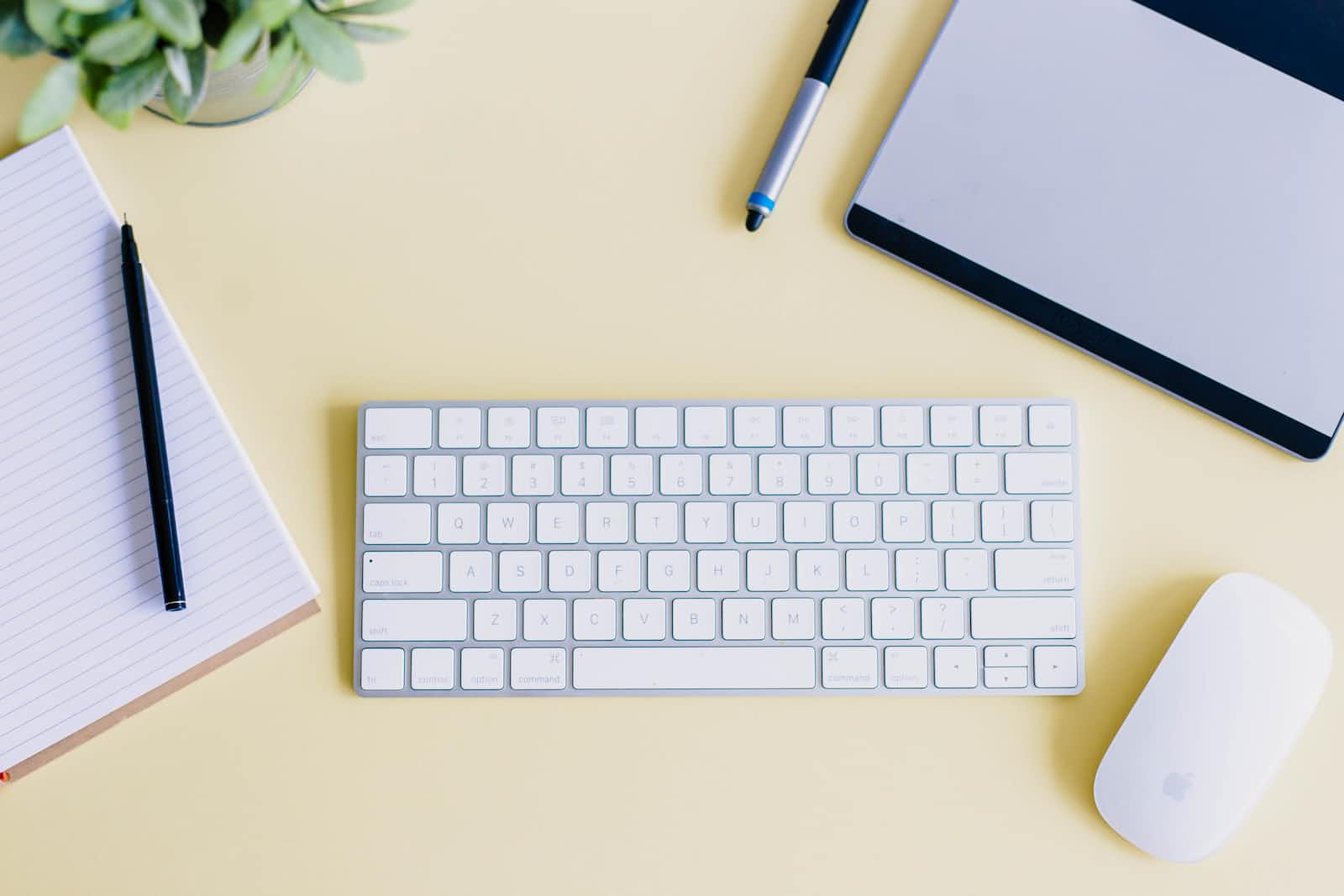 Flat lay of a white keyboard, wireless mouse, notepad with pen, stylus, tablet, and a small plant on a pale yellow desk—perfect for jotting down notes or organizing Reference Stories.