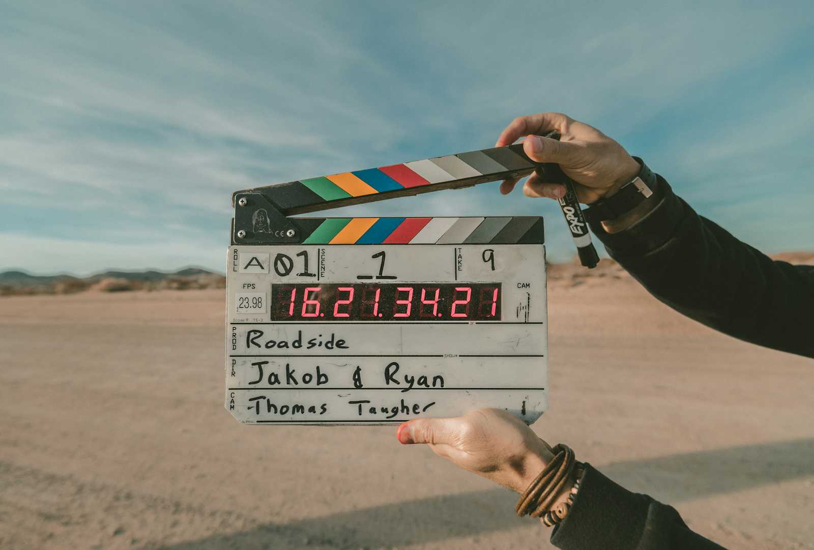 A person holds a film clapperboard in a desert setting, ready to upload videos that capture every scene detail. The clapperboard displays scene information and colorful stripes, with mountains and a blue sky in the background—creating the perfect backdrop for documenting each moment like a Cypress test run.