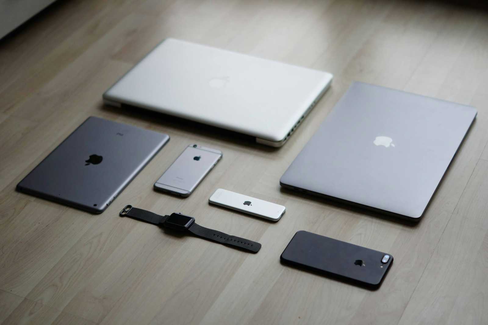 Apple devices including two laptops, an iPad, three iPhones, and a smartwatch are neatly arranged on a light wood floor—perfect for demonstrating cross-platform testing setups.