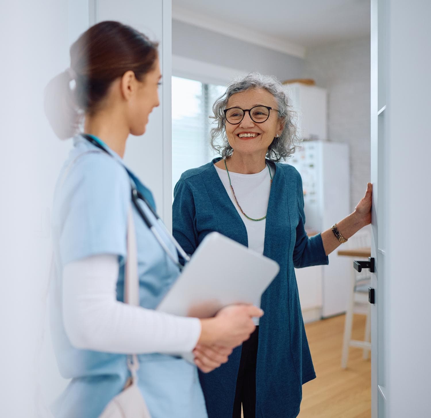 An older woman smiles as she greets a nurse holding a tablet at her door in a bright, modern home setting, highlighting the benefits of an enhanced Digital Patient Experience.